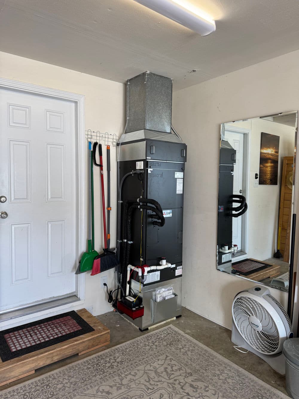 Furnace installed in a garage with cleaning tools, mirror, and a fan nearby.