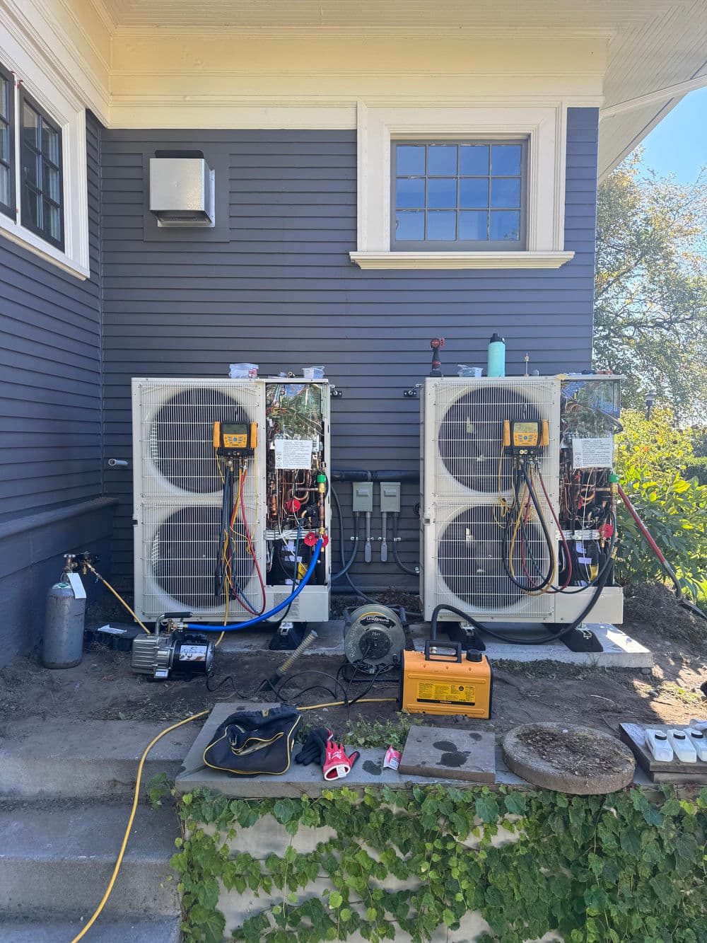 Two air conditioning units installed on a house exterior with tools and equipment nearby.