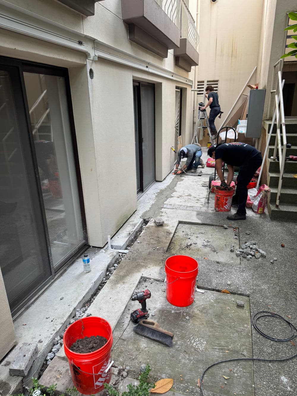 Workers laying concrete and using tools in a construction area outside a building.